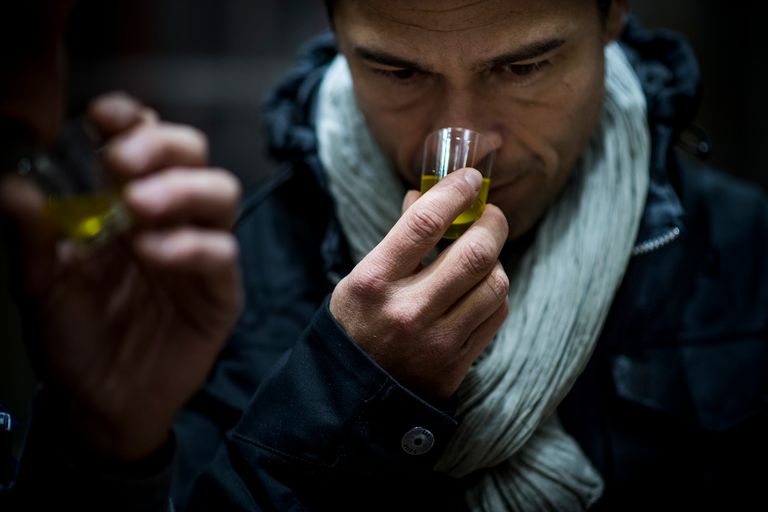 Alexis Muñoz tasting olive oil by smelling its aroma from a small glass, wearing a scarf and a dark jacket, focusing intently on the scent.
