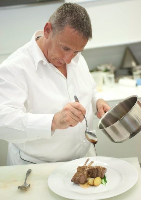 Chez Louis-Michel Lentz habillée d'une veste de cuisinier blanche, en train de dresser une assiette, une cuillière dans sa main, qui fait couler une sauce nappante sur un carré d'agneau.