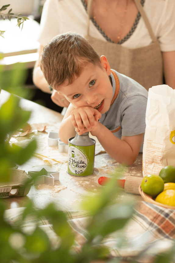 A child holding a can of olive oil for grown ups, Arbequina variety, ripe fruits, produced by Alexis Muñoz, in a kitchen with cookie cutters and flour on the countertop.