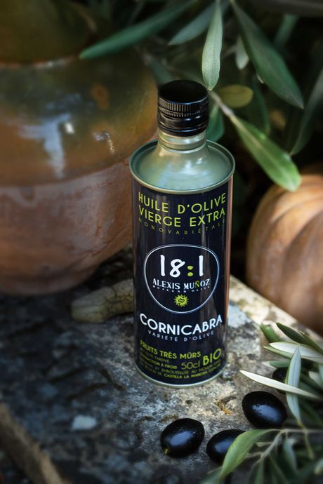 Bidon 500 ml d'huile d'olive, de la marque Alexis Muñoz, de couleur noir, disposé sur une marche en pierre avec un crruchon en terre en arrière plan et en premier plan des branches d'oliviers et des olives noires.