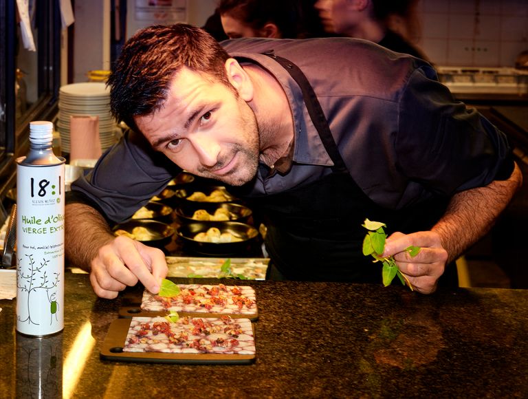 Chef Nicolas Brousse in the kitchen holding aromatic herbs and a can of 18:1 Picual olive oil from the Alexis Muñoz brand.