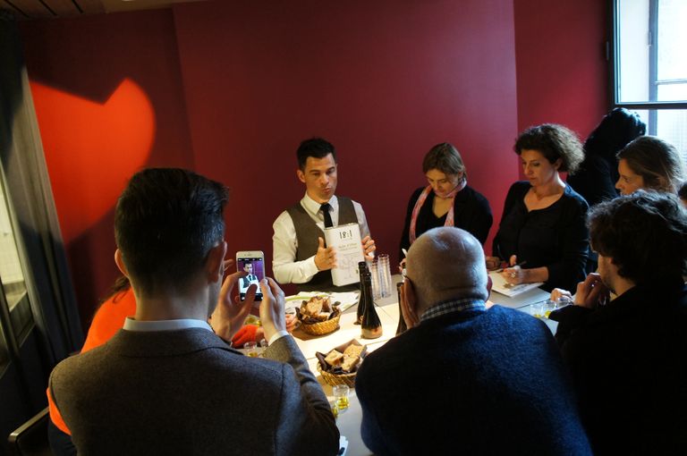 Tasting session with Alexis Muñoz holding a tin of 18:1 extra virgin olive oil, surrounded by an attentive group of people. The setting is a warmly lit room with a red wall, and the table is adorned with bread and olive oil.