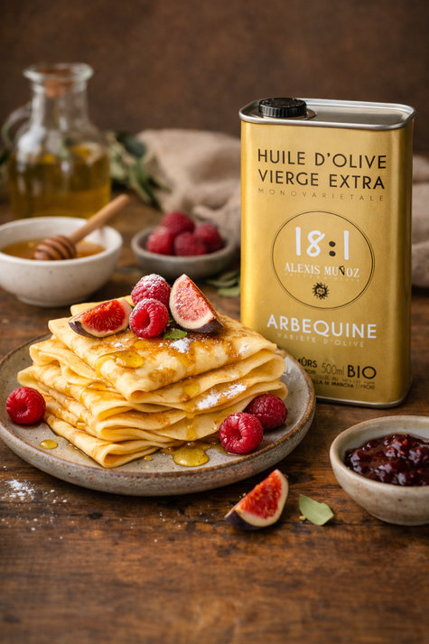 Several crêpes on a grey plate, topped with raspberries, figs, and honey. Three bowls on the side: one with jam, one with honey, and one with raspberries. Behind the crêpes, there is a bottle of 18:1 Arbequina olive oil.