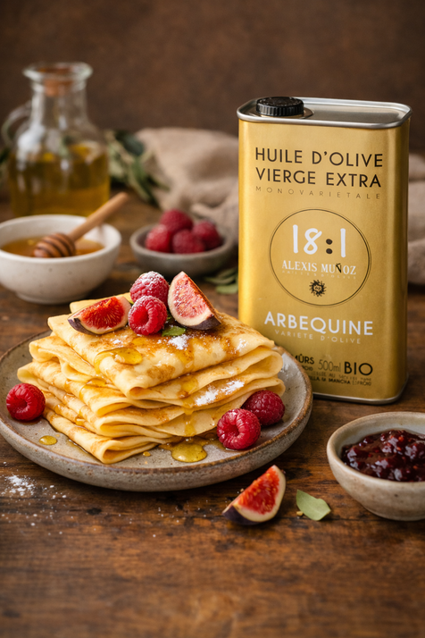 Several crêpes on a grey plate, topped with raspberries, figs, and honey. Three bowls on the side: one with jam, one with honey, and one with raspberries. Behind the crêpes, there is a bottle of 18:1 Arbequina olive oil.