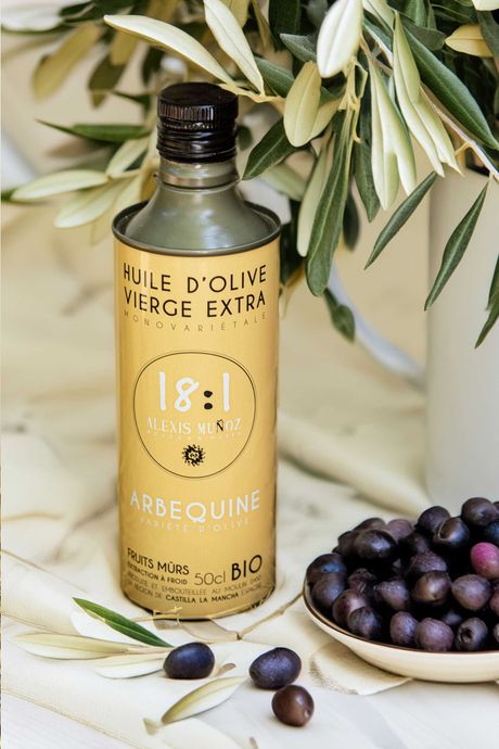 500ml bottle of olive oil, from the brand Alexis Muñoz, in a sandy beige color, placed on a table with a beige tablecloth, with ripe purple olives beside it and a vase with olive branches.