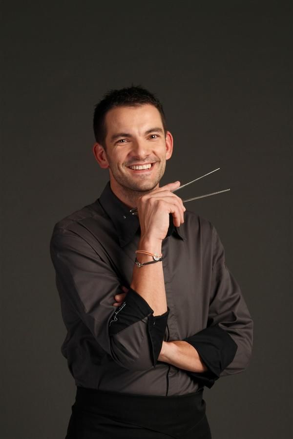 Chef Laurent Peugeot portant une chemise sombre, souriant avec assurance en tenant des baguettes dans une main, les bras partiellement croisés, sur un fond sombre.