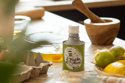 Bidon huile d'olive des futurs grands Arbequina BIO 25cl, sur une table avec en fond un mortier, un bol avec des oeufs, une boîte d'oeufs. Sur la table, il y a de la farine étalée. 