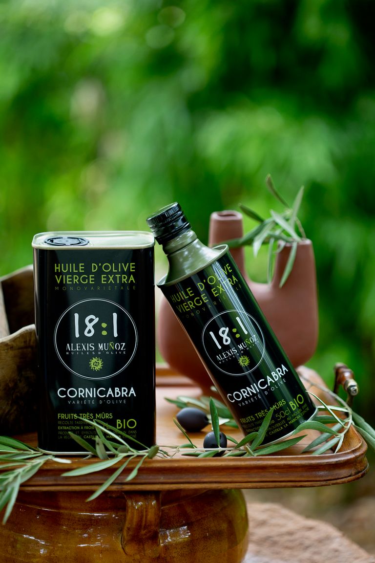 500ml and 1L cans of olive oil from the Alexis Muñoz brand, of the Cornicabra variety. All displayed on a wooden table, with olive branches, black olives, and clay jugs.