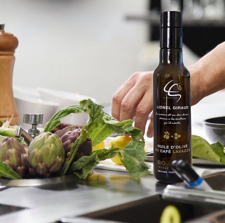 A glass bottle of coffee-infused olive oil, born from the collaboration between Alexis Muñoz, olive oil producer, and Lionel Giraud, Michelin-starred chef, placed on a stainless steel countertop with artichokes and lemons.