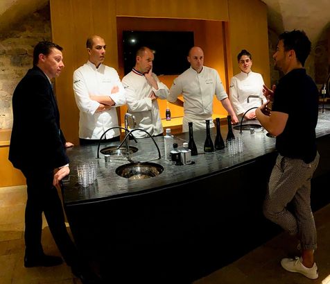 Alexis Muñoz échangeant avec un groupe de chefs cuisiniers autour d'un plan de travail noir, sur lequel sont disposées des bouteilles d'huile d'olive et des verres, dans une salle aux murs en pierre et à l'ambiance chaleureuse.