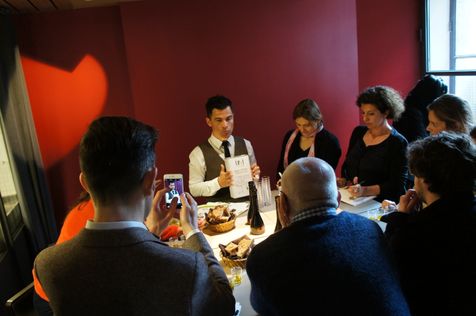 Séance de dégustation avec Alexis Muñoz tenant un bidon d'huile d'olive vierge extra 18:1, entouré d'un groupe de personnes attentives. Le cadre est une pièce chaleureusement éclairée avec un mur rouge, et la table est ornée de pain et d'huile d'olive.