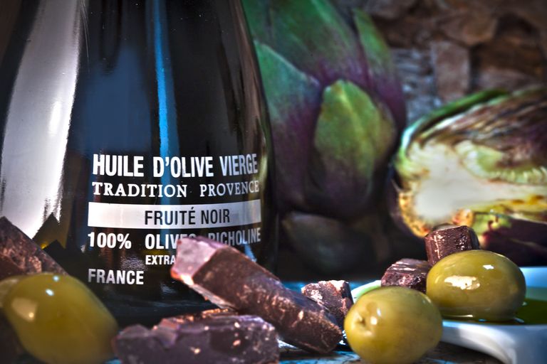 Glass bottle of olive oil from the Alexis Muñoz brand, with chocolate and green olives in the foreground to suggest pairings in pastry.