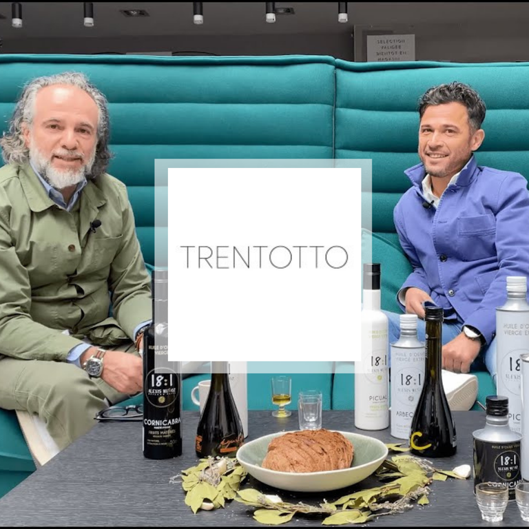 Two men are sitting on a blue sofa. One is wearing a green jacket, the other a blue one. In front of them, there's a table with olive oil, cups, and bread