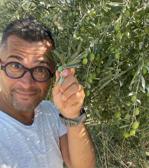 Alexis Muñoz holding an olive branch in his hand, next to an olive tree with branches and green olives. He is wearing glasses, a gray t-shirt, and a watch on his wrist.