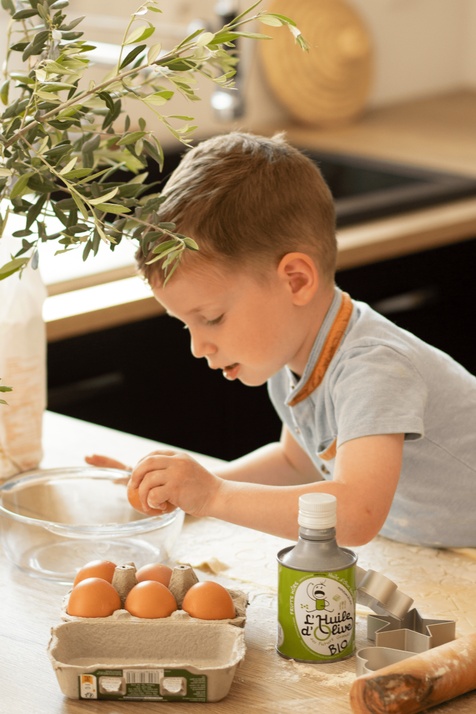 Enfant en train de cuisinner, il casse des oeufs dans un saladier, il porte un tee shirt bleu. A coté il y a un bidon d'huile d'olive des Futurs grands, bidon vert de 25cl