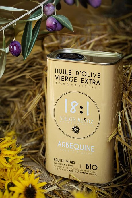 Bidon 1L d'huile d'olive, de la marque Alexis Muñoz, de couleur beige sable, disposé sur un fond avec des olives mûres, du foin et des tournesols jaunes.