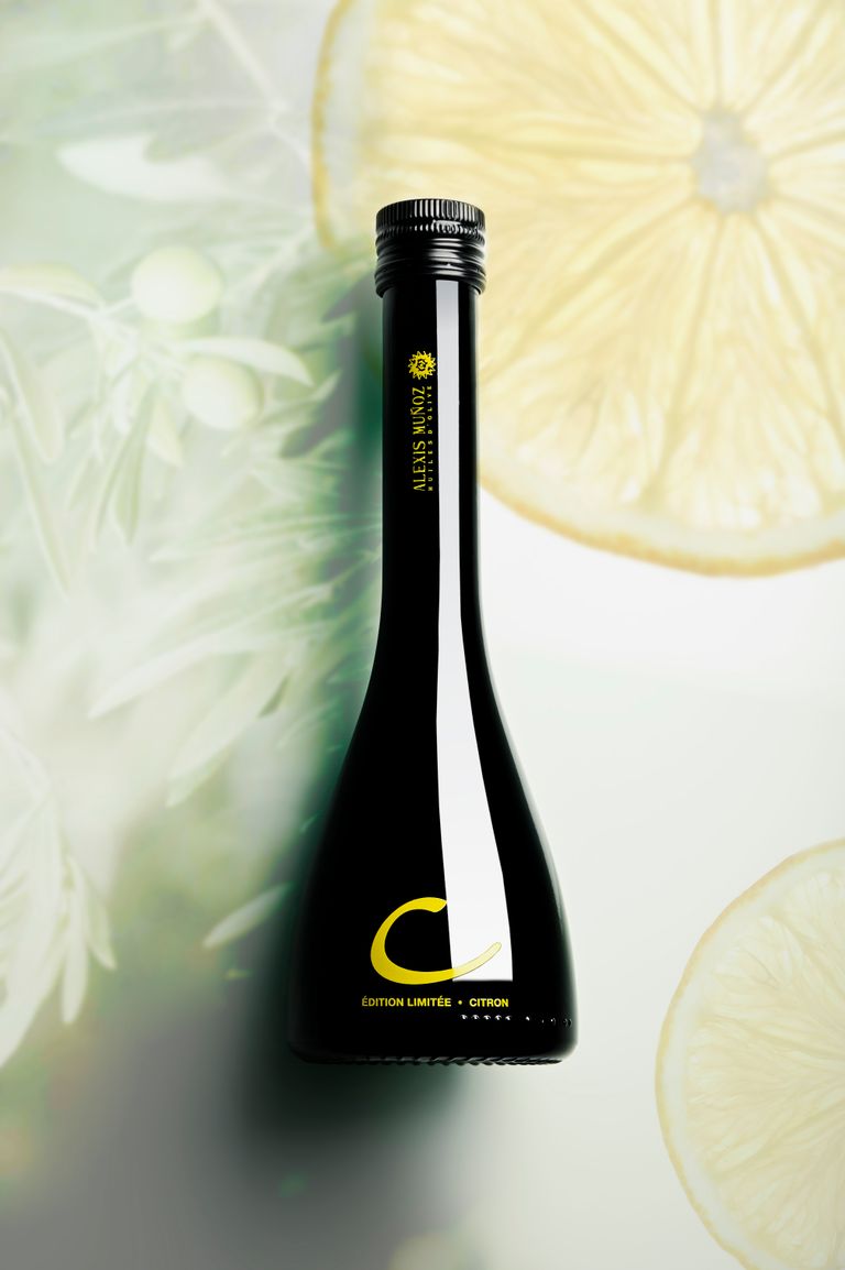 250ml glass bottle of olive oil and lemon pressed together, limited edition, from the Alexis Muñoz brand. The bottle is placed on a background with olive branches and cross-sectioned lemons.