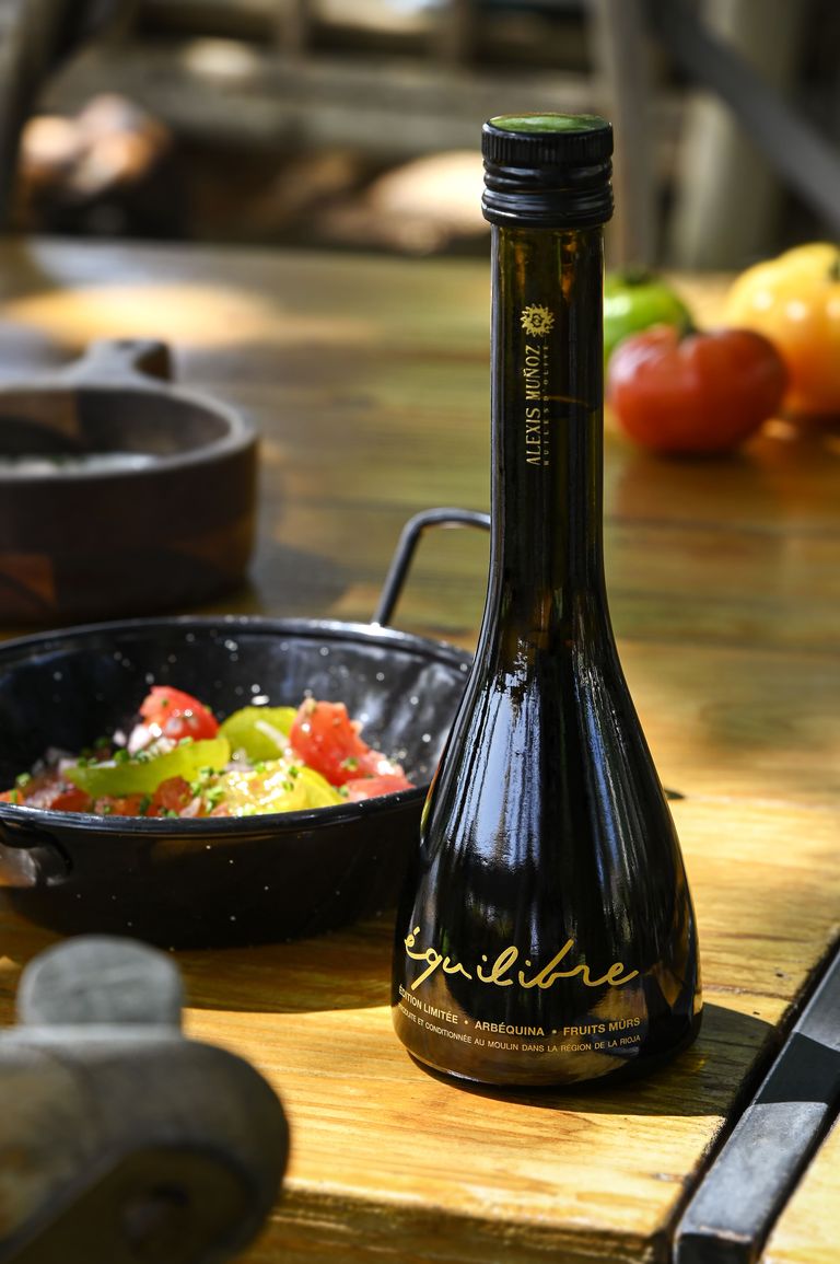 250ml bottle of Limited Edition Equilibre olive oil from the Alexis Muñoz brand. The bottle is placed on a wooden table with tomatoes in the background and a tomato salad to its left, accompanied by aromatic herbs.