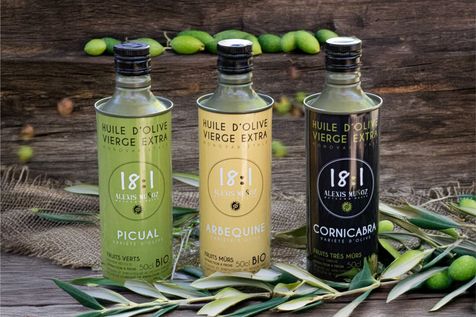 3 bouteilles d'huiles d'olives 18.1, variétés Picual, Arbequine, Cornicabra en 50 cl. Posées sur une branche et des feuilles d'olivier. Olives vertes en fond sur un support en bois. 