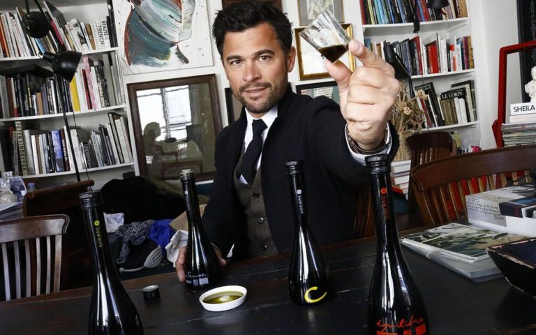 Alexis Muñoz sitting at a table surrounded by books, holding a glass of olive oil in his hand, with several glass bottles of his Limited Edition olive oils in front of him.