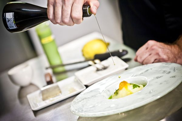 Kitchen scene. A chef is preparing a dish, drizzling a stream of 18:1 olive oil into a shallow bowl filled with mashed potatoes. In the background: the stainless-steel countertop of a professional kitchen, with plates, a yellow lemon, and various cooking