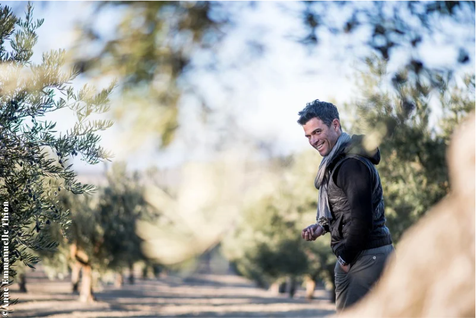 Alexis Muñoz souriant dans les allées d'une oliveraie en arrière plan