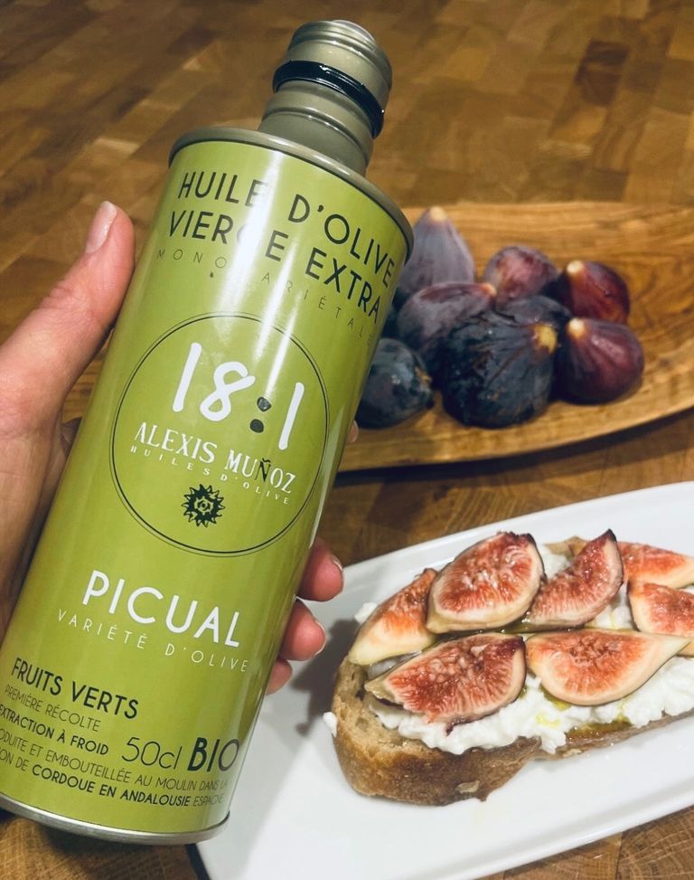 A bottle of Picual olive oil (18:1) in green, held in the left hand. In the background, a plate with a slice of bread topped with creamy burrata and sliced figs. A wooden board with whole figs. Everything arranged on a wooden table.