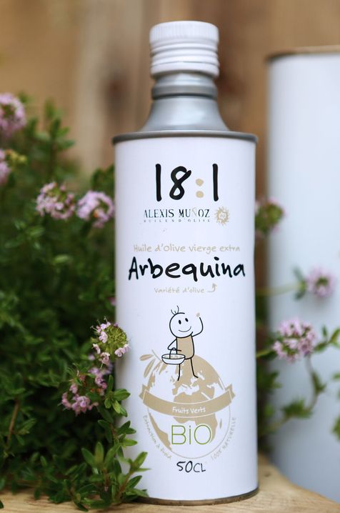 Photo d'ambiance d'un bidon cylindrique blanc d'huile d'olive variété Arbequina, de la marque Alexis Muñoz 18:1? avec des fleurs champêtres rose et verte en arrière plan. 