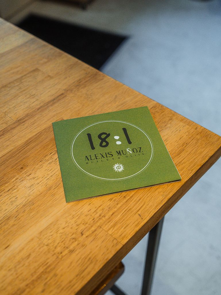 Dépliant vert format carré, de la marque d'huile d'olive 18:1 Alexis Muñoz, de la variété Arbequina et Cornicabra, posée sur une table de restaurant en bois avec des verres à pieds.