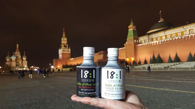 Bidons d'huile d'olive cylindriques blanc et noir de la marque Alexis Muñoz, 18:1, posées sur une main, avec en fond la place Rouge de Moscou, en Russie.