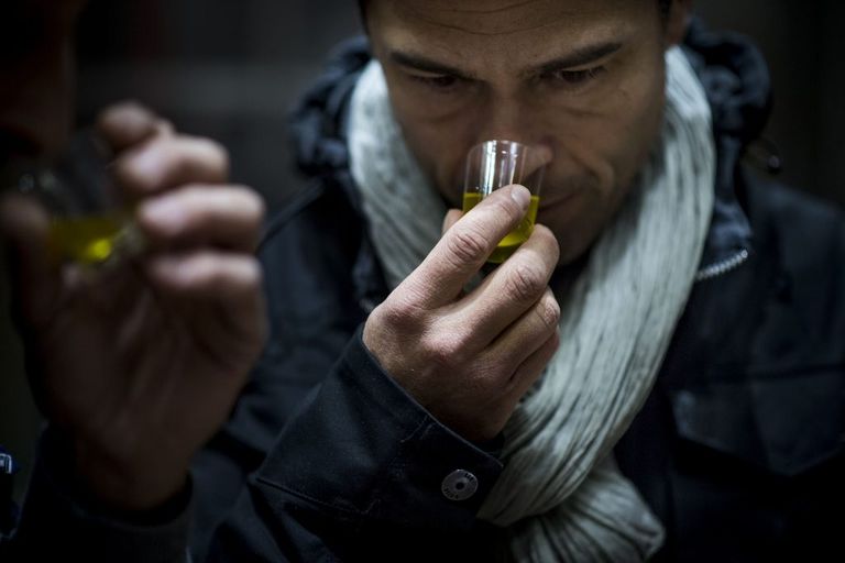 A man holds a cup in his right hand. Inside the cup, there is olive oil. The man is smelling the olive oil.