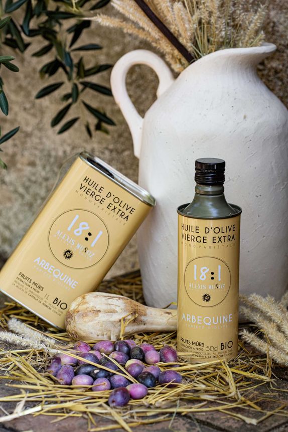 Bidons 500ml et 1L d'huile d'olive, de la marque Alexis Muñoz, de couleur beige sable, disposé sur un fond avec un cruchon blanc cassé, des épis de blé et des olives mûres.