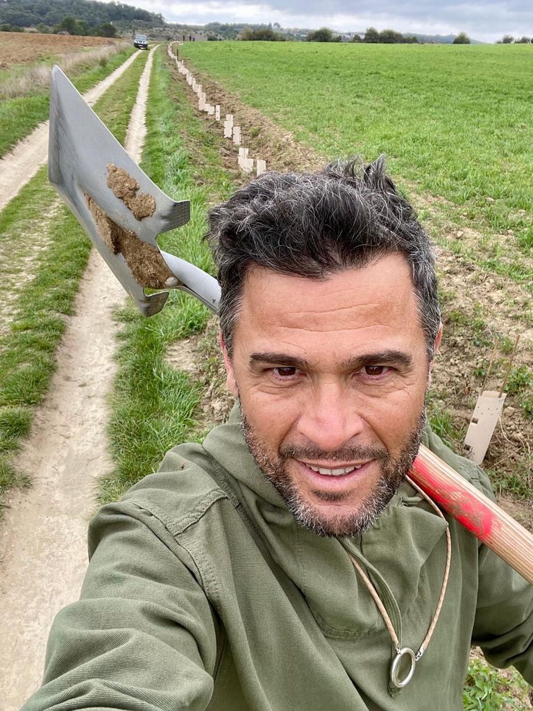 Alexis Muñoz with a pickaxe on his shoulder, standing in an olive grove.