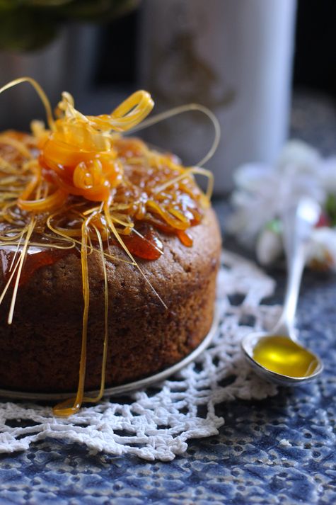 Chestnut cake topped with a caramel glaze. Presented on a white doily over a blue tablecloth, with a teaspoon of olive oil on the side. In the background, a white bottle of Alexis Muñoz Arbequina olive oil.