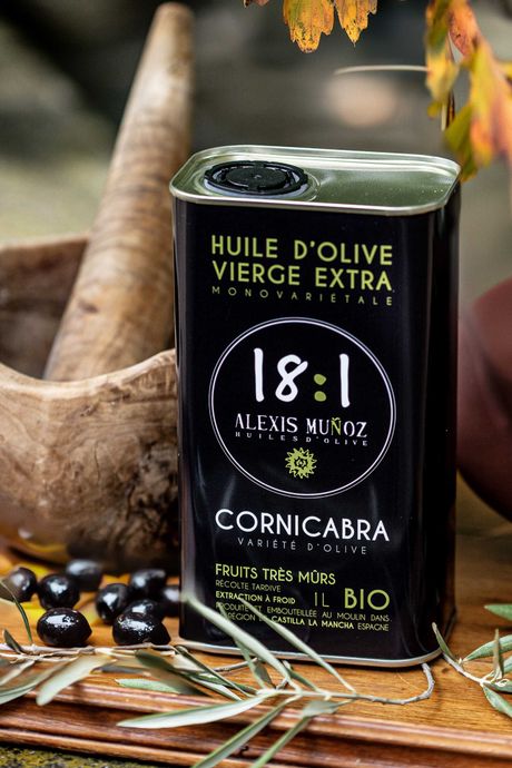 1L can of olive oil, from the brand Alexis Muñoz, in a black color, placed on a wooden table, with a mortar in the background, and olive branches and black olives in the foreground.