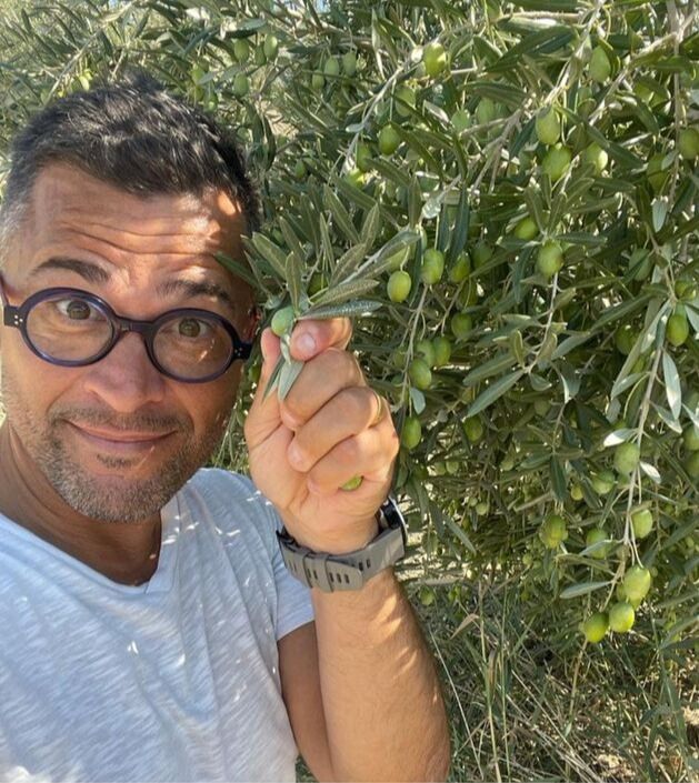 Alexis Muñoz tenant une branche d'olivier dans sa main, à côté d'un olivier avec des branches et des olives vertes. il porte des lunettes, un t shirt gris et une montre a son poignet