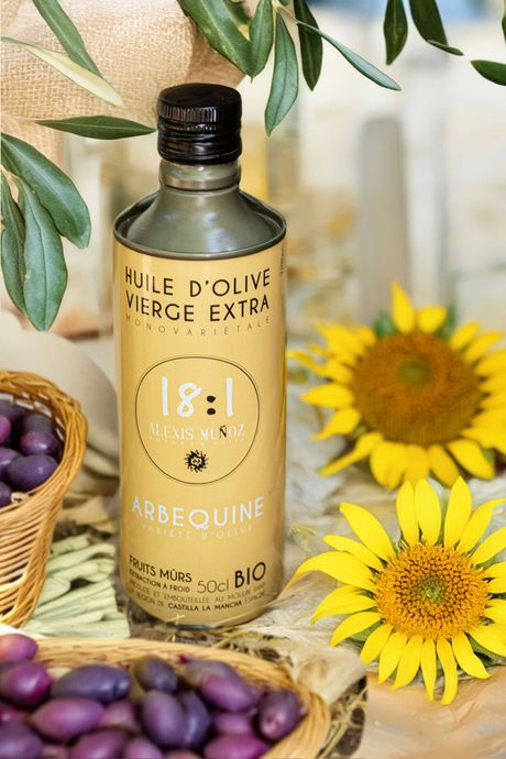 A 50cl tin of 18.1 Arbequina olive oil, surrounded by sunflower blossoms and olive branches, placed in a wicker basket filled with ripe olives, set on a white table.