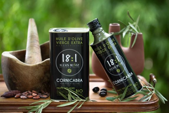 500ml and 1L cans of olive oil from the Alexis Muñoz brand, black in color, arranged on a background with earthenware jugs, olive branches, and very ripe black olives.