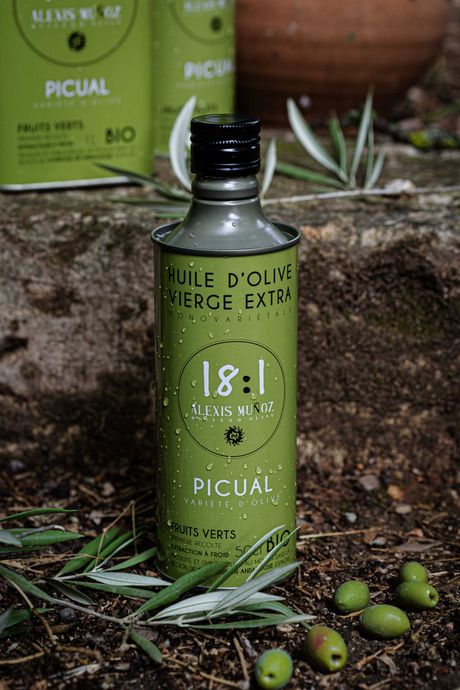 500ml bottle of olive oil, from the brand Alexis Muñoz, in a green color, on a stone background, with olive branches and green olives on the ground.