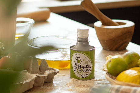 Bidon huile d'olive des futurs grands Arbequina BIO 25cl, sur une table avec en fond un mortier, un bol avec des oeufs, une boîte d'oeufs. Sur la table, il y a de la farine étalée.
